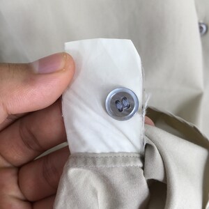Peut inclure: Gros plan sur un d&eacute;tail de chemise beige boutonn&eacute;e. Un bouton bleu clair est cousu sur une patte de tissu blanc, le tissu beige de la chemise &eacute;tant visible en dessous. L'image met en &eacute;vidence le bouton et la confection de la chemise.