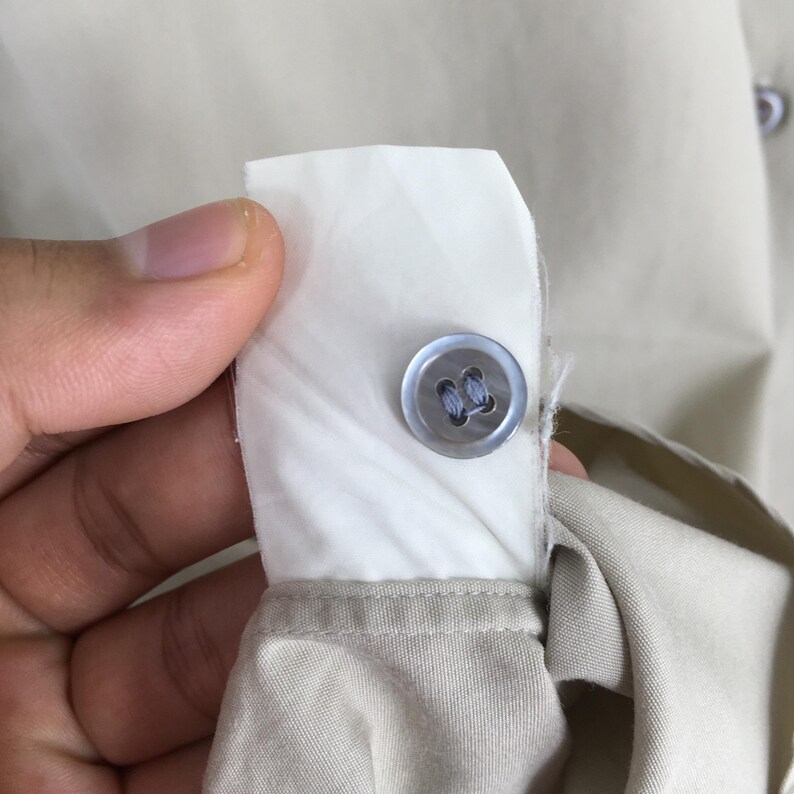 Peut inclure: Gros plan sur un d&eacute;tail de chemise beige boutonn&eacute;e. Un bouton bleu clair est cousu sur une patte de tissu blanc, le tissu beige de la chemise &eacute;tant visible en dessous. L'image met en &eacute;vidence le bouton et la confection de la chemise.