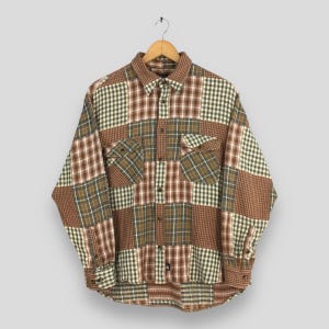 May include: A button-down shirt with a patchwork design, featuring various plaid patterns in brown, green, and white. It has two chest pockets and buttoned cuffs. The shirt is displayed on a wooden hanger. The shirt's style is casual.