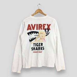 May include: White long-sleeve shirt with a graphic on the back featuring the word "AVIREX" in red, a shark mouth design, and the words "TIGER SHARKS". The shirt also has a graphic of a woman in a tutu.