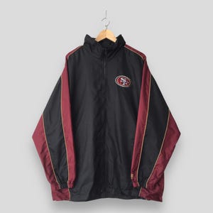Könnte beinhalten: Eine schwarze Windjacke mit kastanienbraunen Seiteneinsätzen und dünnen goldenen Paspeln. Die Jacke hat einen hohen Kragen, einen durchgehenden Frontreißverschluss und ein San Francisco 49ers Logo-Emblem auf der linken Brustseite.