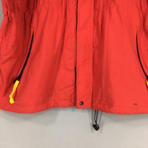 May include: Vibrant red jacket featuring black zips and yellow pull tabs. The jacket includes a drawstring at the hem and a button fastening. It seems to be crafted from a lightweight, water-resistant fabric.