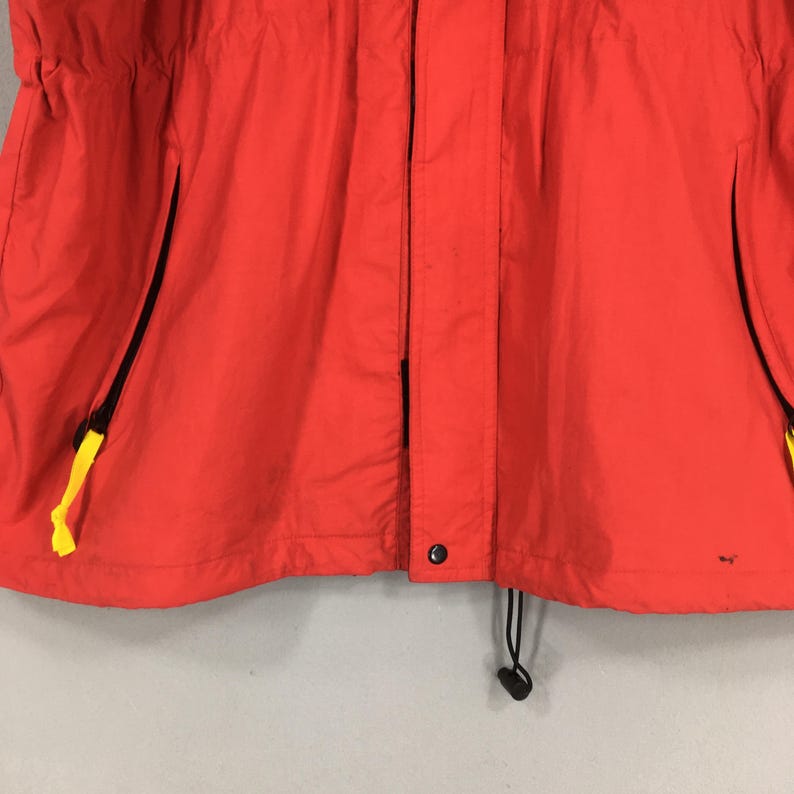 May include: Vibrant red jacket featuring black zips and yellow pull tabs. The jacket includes a drawstring at the hem and a button fastening. It seems to be crafted from a lightweight, water-resistant fabric.