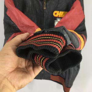 May include: Black and red leather jacket with the word "CHIEFS" in yellow. The jacket has red sleeves and a black ribbed cuff with red and yellow stripes. The jacket is partially zipped.