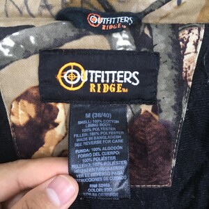 May include: Close-up of a jacket tag with the "Outfitters Ridge" logo in orange and white. The tag includes size M (38/40) and fabric composition details: shell 100% cotton, lining 100% polyester, and filler 100% polyester.