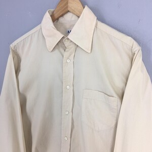 Pode incluir: Uma camisa de bot&atilde;o de manga comprida, de cor creme, com um bolso no peito. A camisa tem um colarinho pontudo e uma frente de bot&atilde;o.