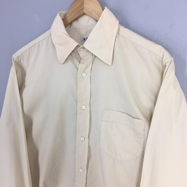 Pode incluir: Uma camisa de bot&atilde;o de manga comprida, de cor creme, com um bolso no peito. A camisa tem um colarinho pontudo e uma frente de bot&atilde;o.