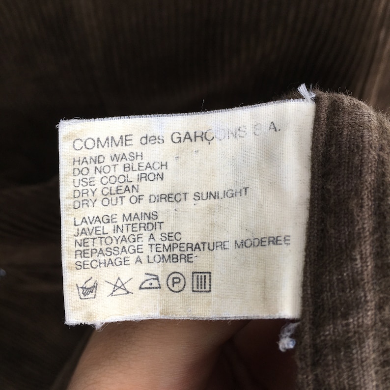 Puede incluir: Primer plano de una etiqueta de tela marr&oacute;n con el texto "COMME des GAR&Ccedil;ONS 6 A." e instrucciones de cuidado en ingl&eacute;s y franc&eacute;s. La etiqueta est&aacute; adherida a una prenda de pana marr&oacute;n.