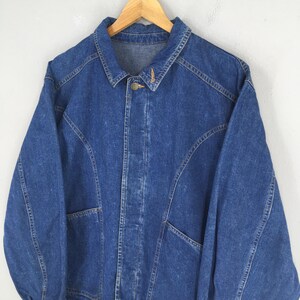 May include: A vintage blue denim jacket with a button-up front and two front pockets. The jacket is hanging on a wooden hanger against a plain white background. The jacket has a classic collar and long sleeves.