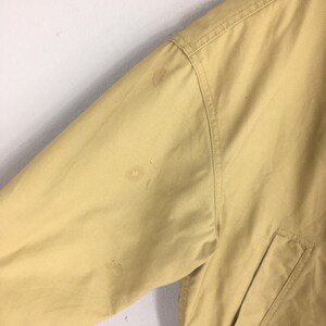 May include: Close-up of a tan jacket sleeve with a pocket and visible stains. The jacket appears to be made of a durable fabric, with seams and stitching details. The sleeve is angled, suggesting a casual or workwear style.