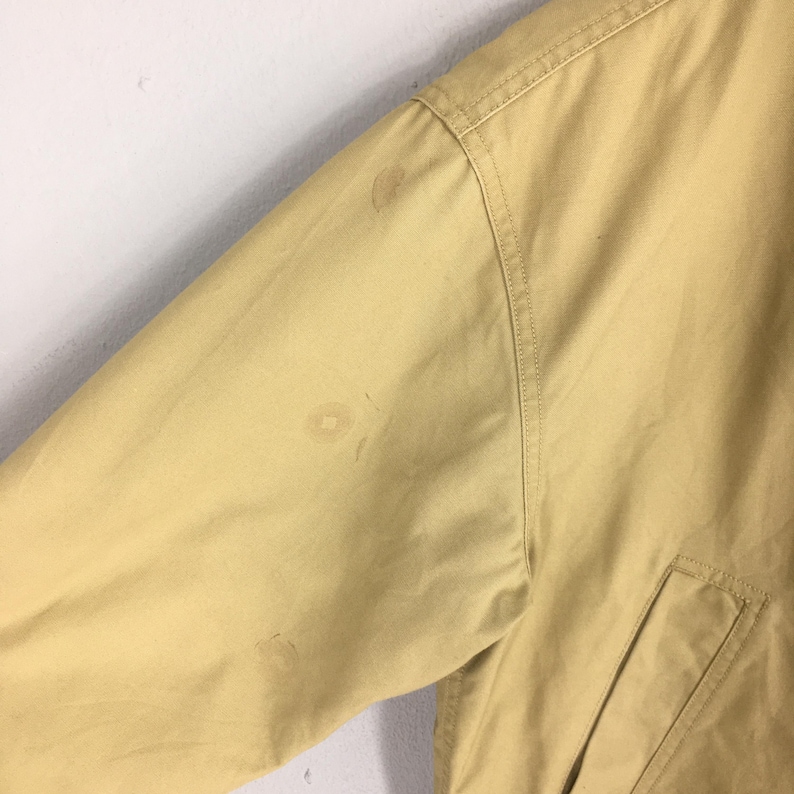 May include: Close-up of a tan jacket sleeve with a pocket and visible stains. The jacket appears to be made of a durable fabric, with seams and stitching details. The sleeve is angled, suggesting a casual or workwear style.