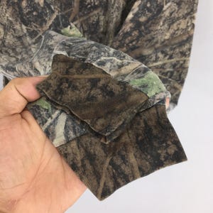 May include: Long-sleeved camouflage shirt. The shirt features a brown, green, and tan pattern designed to blend with a forest environment. The cuff of the sleeve is visible, showing the fabric's texture and color variations.