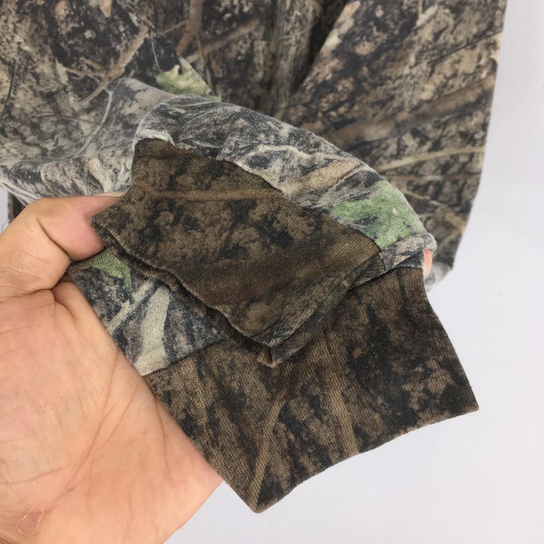 May include: Long-sleeved camouflage shirt. The shirt features a brown, green, and tan pattern designed to blend with a forest environment. The cuff of the sleeve is visible, showing the fabric's texture and color variations.