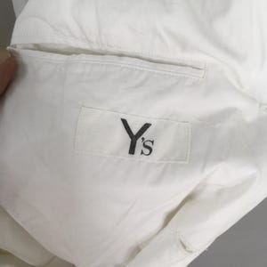 May include: A white garment featuring a rectangular fabric label stitched onto the interior lining. The label displays the black text Y's in a serif font. The garment includes a horizontal welt pocket opening positioned above the brand label.