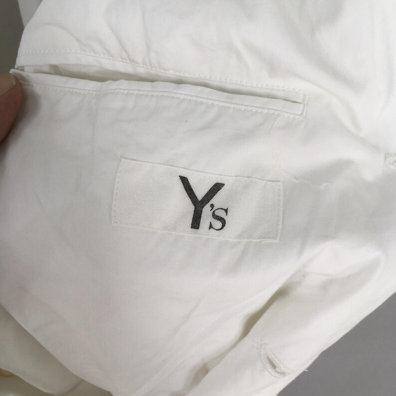 May include: A white garment featuring a rectangular fabric label stitched onto the interior lining. The label displays the black text Y's in a serif font. The garment includes a horizontal welt pocket opening positioned above the brand label.