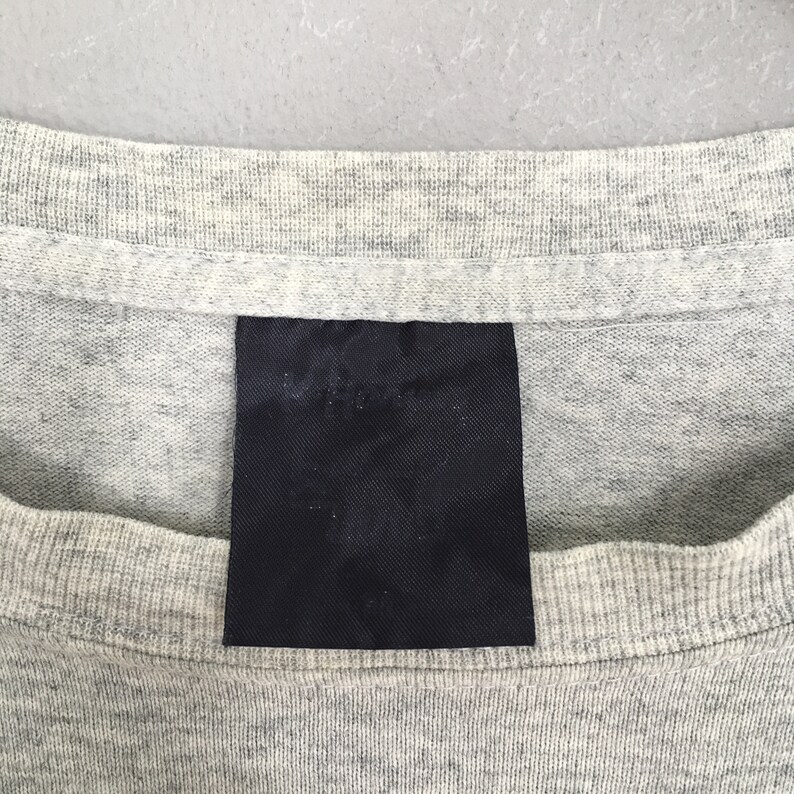 May include: Close-up of a gray t-shirt with a black fabric label sewn on the front. The label is blank.