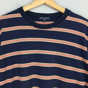 Puede incluir: Camiseta a rayas azul marino con rayas horizontales en beige y rojo. La camiseta tiene cuello redondo y mangas cortas. La marca "COMME des GARCONS HOMME" es visible en la etiqueta.