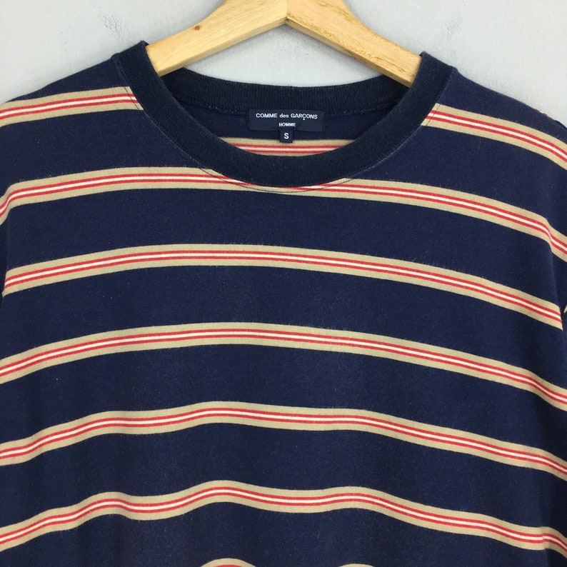 Puede incluir: Camiseta a rayas azul marino con rayas horizontales en beige y rojo. La camiseta tiene cuello redondo y mangas cortas. La marca "COMME des GARCONS HOMME" es visible en la etiqueta.