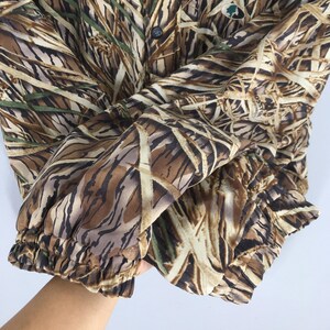 Puede incluir: Chaqueta de camuflaje con un patr&oacute;n marr&oacute;n, beige y verde que imita juncos y hierbas. La chaqueta tiene cierre de botones y pu&ntilde;os el&aacute;sticos. El logotipo de Mossy Oak es visible en la parte superior derecha.