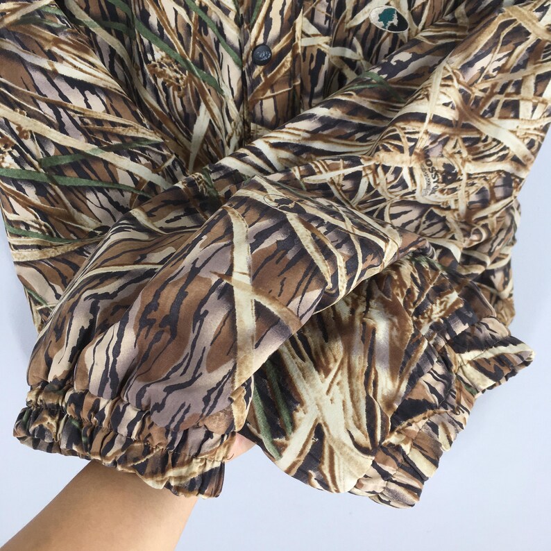 Puede incluir: Chaqueta de camuflaje con un patr&oacute;n marr&oacute;n, beige y verde que imita juncos y hierbas. La chaqueta tiene cierre de botones y pu&ntilde;os el&aacute;sticos. El logotipo de Mossy Oak es visible en la parte superior derecha.