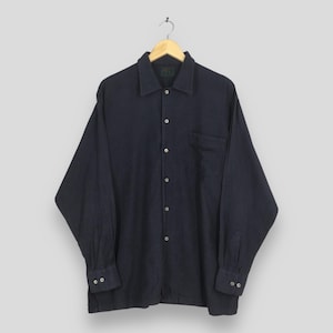 Puede incluir: Una camisa azul marino de manga larga con botones, un bolsillo en el pecho y pu&ntilde;os abotonados. La camisa est&aacute; colgada de una percha de madera sobre un fondo gris claro. La camisa tiene un cuello cl&aacute;sico.