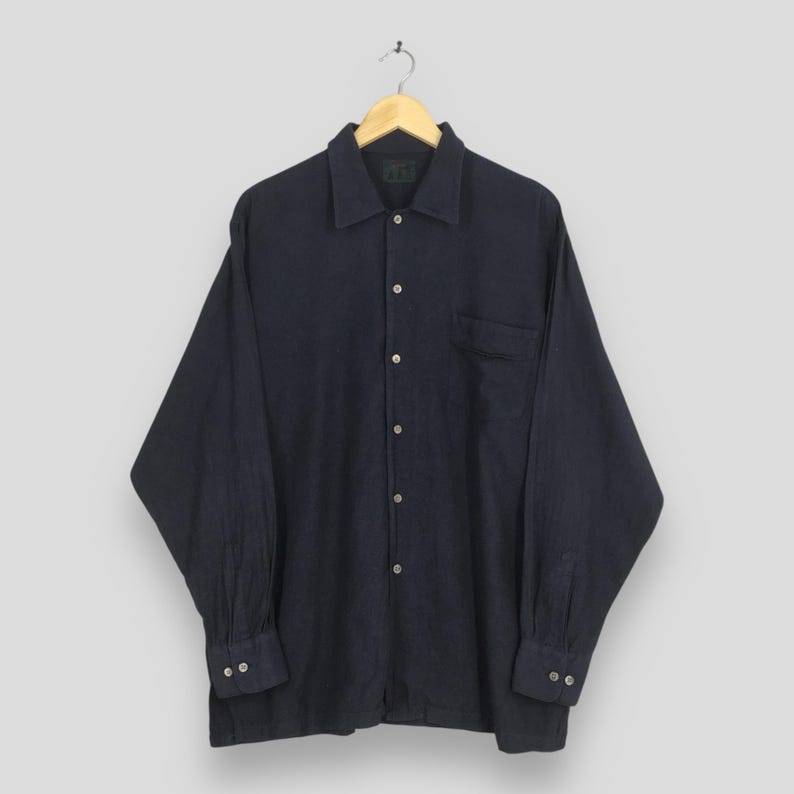 Puede incluir: Una camisa azul marino de manga larga con botones, un bolsillo en el pecho y pu&ntilde;os abotonados. La camisa est&aacute; colgada de una percha de madera sobre un fondo gris claro. La camisa tiene un cuello cl&aacute;sico.