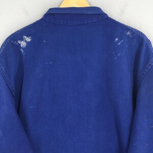Peut inclure: Une veste de travail bleu roi avec un col boutonn&eacute; et des manches longues. Le dos de la veste pr&eacute;sente des &eacute;claboussures de peinture blanche. La veste est suspendue &agrave; un cintre en bois.