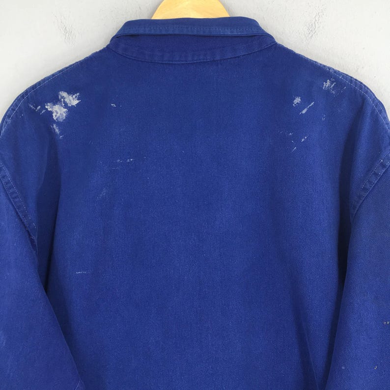 Peut inclure: Une veste de travail bleu roi avec un col boutonn&eacute; et des manches longues. Le dos de la veste pr&eacute;sente des &eacute;claboussures de peinture blanche. La veste est suspendue &agrave; un cintre en bois.