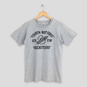 May include: A heather gray t-shirt with a black graphic featuring a rocket and the text "FOURTH BUT FIRST" and "ROCKETEERS". The shirt is hanging on a wooden hanger.