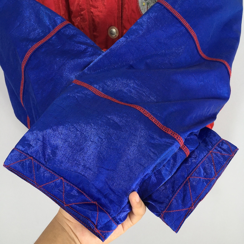 May include: Close-up of a vintage jacket with a color-blocked design. The jacket features a red upper section and blue sleeves with red stitching. The sleeves are folded, showcasing the vibrant colors and textures of the fabric. The jacket has a button closure.