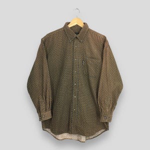 Puede incluir: Camisa abotonada marrón con un pequeño patrón repetitivo de cuadrados blancos y amarillos. La camisa de manga larga tiene cuello abotonado, bolsillo en el pecho y puños abotonados. La camisa está colgada de una percha de madera.