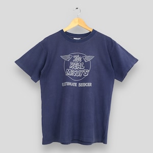 May include: A navy blue short-sleeve t-shirt featuring a circular graphic on the chest. The graphic includes a pair of wings, the text "The Real McCoy's" in a stylized font, and the words "ULTIMATE SEEKER" printed in capital letters below.