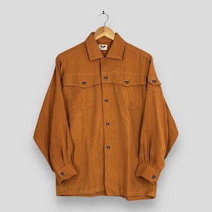 May include: A long-sleeved, rust-colored shirt with a collared neckline and button-down closure. The shirt features two chest pockets and buttoned cuffs. The garment is hanging on a wooden hanger.