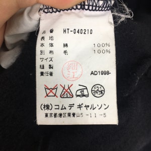 Puede incluir: Una etiqueta blanca con texto negro y s&iacute;mbolos. La etiqueta dice "HT-040210", "100% algod&oacute;n", "100% lana", "AD1998-" y "(株)コムデギャルソン" (Komdegyaruso). La etiqueta tambi&eacute;n tiene una etiqueta de cuidado con s&iacute;mbolos para lavar, blanquear, planchar y secar.