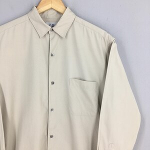 Peut inclure: Chemise boutonn&eacute;e beige clair avec un col et une poche poitrine unique. La chemise a des manches longues et un design classique. Les boutons sont de la m&ecirc;me couleur que la chemise. La chemise est suspendue &agrave; un cintre en bois.