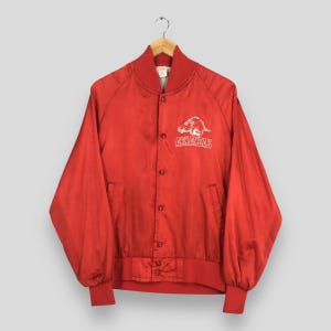 May include: A bright red long-sleeve button-up jacket featuring a white graphic of a razorback hog and the text ARKANSAS printed on the upper left chest. The jacket has a ribbed collar, cuffs, and hem, with a smooth finish and snap button closure.