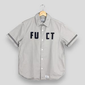 May include: A light gray short-sleeved button-down shirt with the letters "FU CT" appliqued on the front. The shirt has black and white striped accents on the sleeves and is hanging from a wooden hanger.