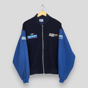 Puede incluir: Chaqueta azul marino con cremallera y mangas azules. La chaqueta presenta parches con el texto "FORMULA", "MILD SEVEN" y "Benetton Formula 1". Las mangas son de color azul liso. La chaqueta está colgada en una percha de madera.