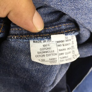 May include: Close-up of a blue denim garment tag. The tag is white with black text, including "MADE IN ITA" and washing instructions. The fabric is a dark blue denim.