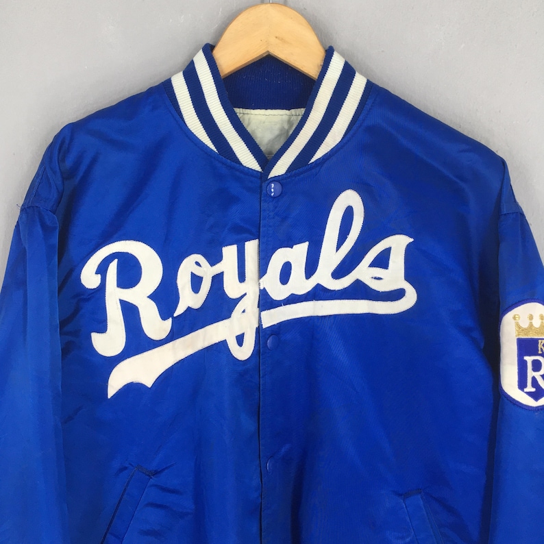 May include: A royal blue satin varsity jacket featuring the word Royals in white script across the chest. The jacket has a ribbed collar with white stripes, a snap button front closure, and a white circular patch on the sleeve with a gold crown.