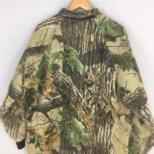 May include: A camouflage jacket with a tree bark and leaf design in brown, green, and tan. The jacket has a collared neckline and brown cuffs. The pattern is designed to blend with natural surroundings. The jacket is designed for outdoor activities.
