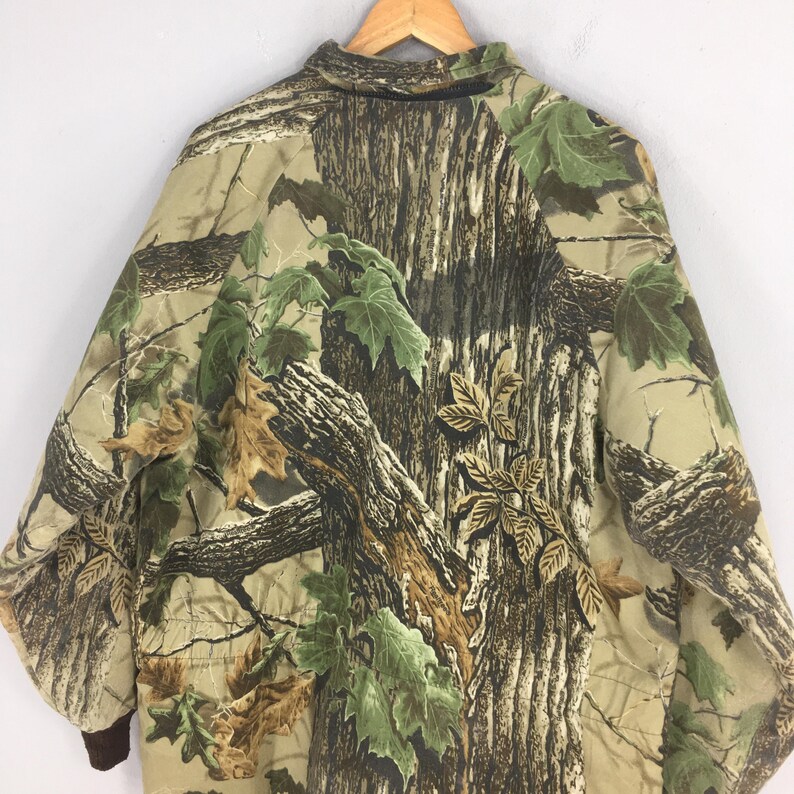 May include: A camouflage jacket with a tree bark and leaf design in brown, green, and tan. The jacket has a collared neckline and brown cuffs. The pattern is designed to blend with natural surroundings. The jacket is designed for outdoor activities.