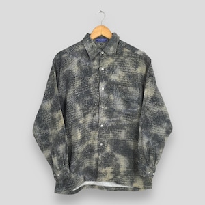 Puede incluir: Una camisa abotonada de manga larga con un patrón texturizado en tonos grises y beige. La camisa tiene un bolsillo en el pecho y está colgada de una percha de madera.