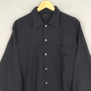 Puede incluir: Una camisa azul marino oscuro de manga larga con botones en la parte delantera y un bolsillo en el pecho. La camisa tiene cuello y est&aacute; colgada de una percha de madera. La marca "A.A.R." es visible en una peque&ntilde;a etiqueta en el cuello.