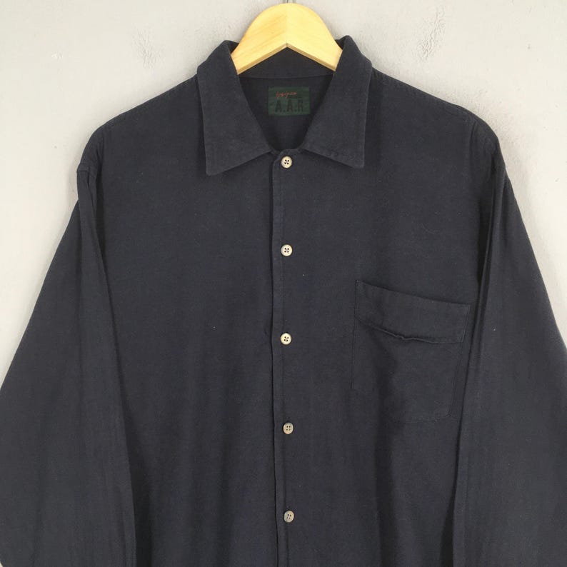 Puede incluir: Una camisa azul marino oscuro de manga larga con botones en la parte delantera y un bolsillo en el pecho. La camisa tiene cuello y est&aacute; colgada de una percha de madera. La marca "A.A.R." es visible en una peque&ntilde;a etiqueta en el cuello.