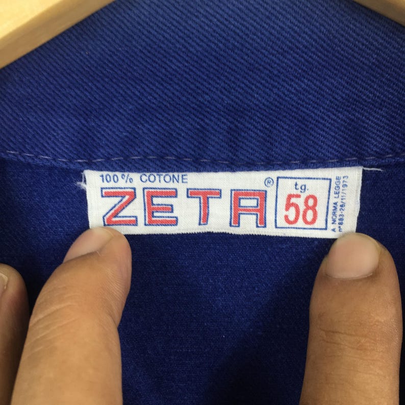 Peut inclure: Gros plan sur un v&ecirc;tement bleu avec une &eacute;tiquette blanche. L'&eacute;tiquette indique "100% COTONE" et "ZETA" avec la taille "58". Le texte suppl&eacute;mentaire comprend "A NORMA LEGGE" et "883-26/11/1973".