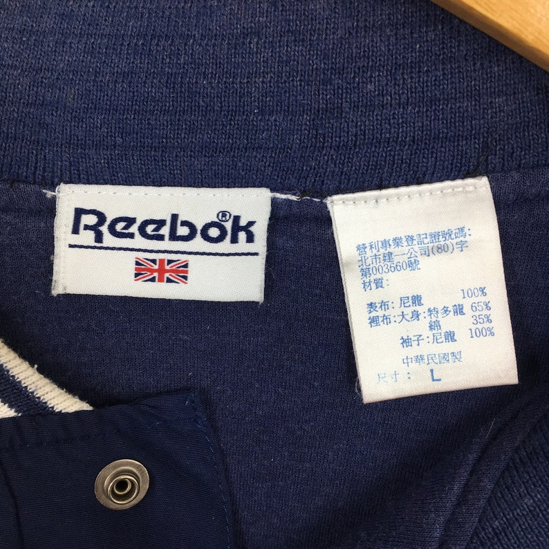 May include: A navy blue Reebok jacket with a white and navy blue striped collar. The jacket has a zipper closure and a button on the left side. The tag on the jacket says "Reebok" with a Union Jack flag and lists the materials and size as "L".