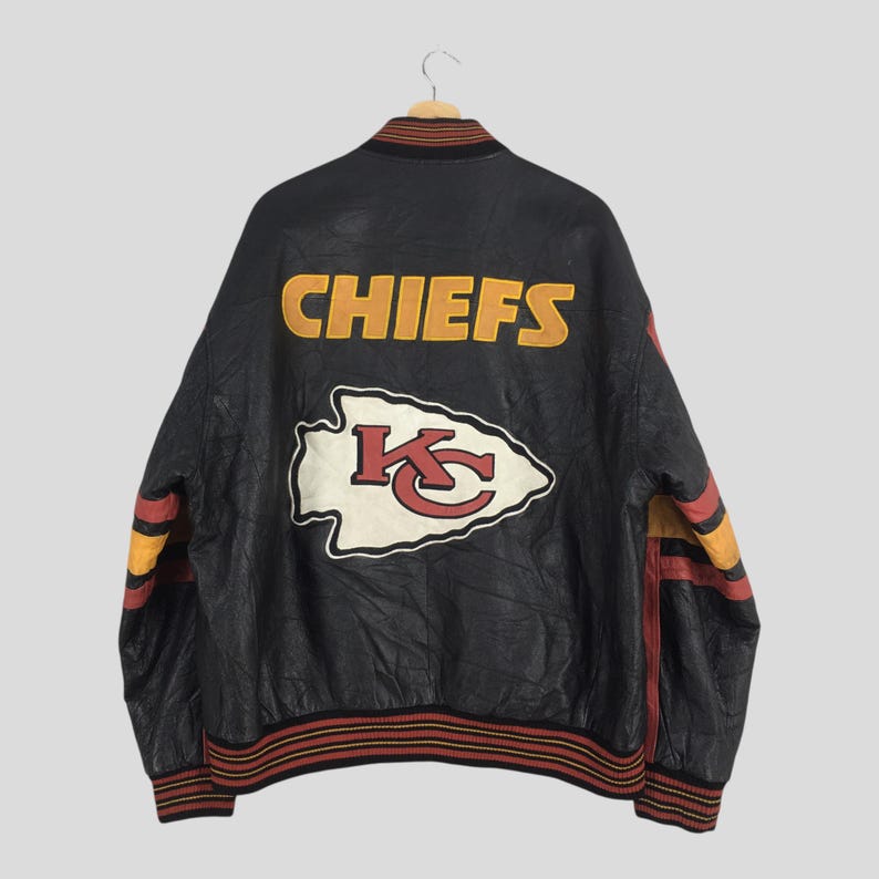May include: Black leather jacket with the word "CHIEFS" in yellow block letters across the back. The jacket features a white and red arrowhead logo and red, yellow, and black striped accents on the sleeves and collar.
