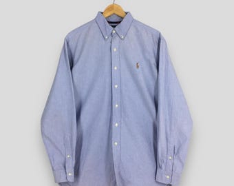 May include: A light blue button-down shirt with long sleeves, hanging on a wooden hanger. The shirt features a small embroidered emblem on the chest and white buttons. The fabric appears to be a soft, woven material.