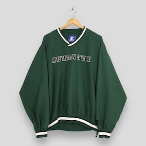 Puede incluir: Un jersey verde de Michigan State con cuello en V blanco y ribete a rayas blancas y negras en la cintura y los puños. Las palabras "MICHIGAN STATE" están bordadas en el pecho con letras blancas.